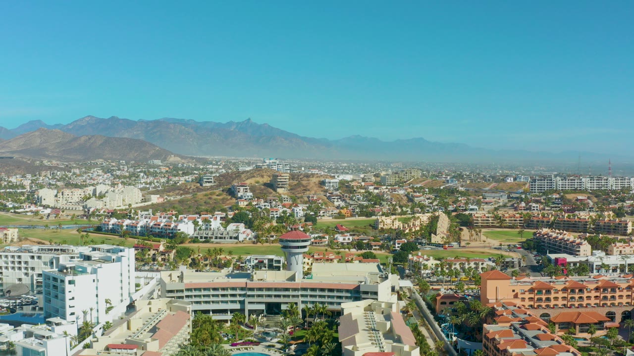Early frame of San Jose Del Cabo from above, with white buildings in the foreground, a red-topped control tower near center, distant mountains, clear blue sky, and no visible people.