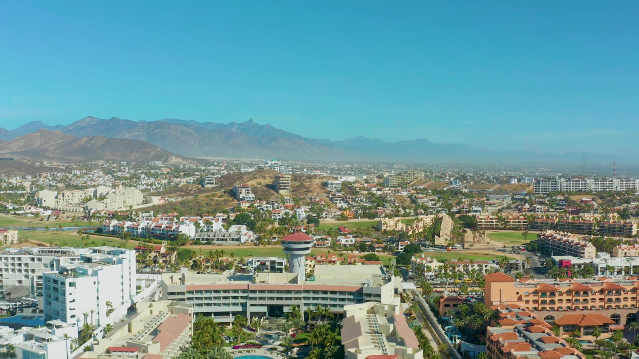 San Jose Del Cabo aerial view one second in, showing resort-style buildings, green areas, a control tower slightly right of center, hazy mountains, and no visible people.