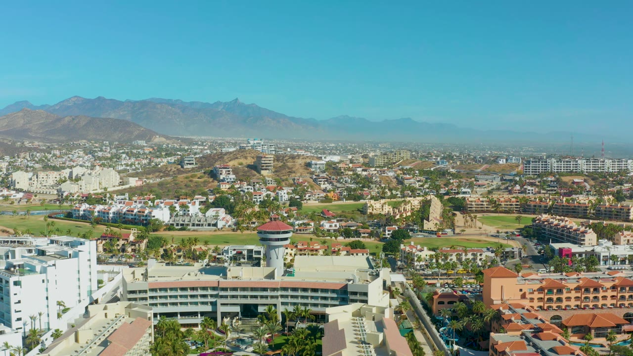 Elevated San Jose Del Cabo city view with low-rise white buildings in front, terracotta-roofed structures to the right, green space through the center, mountains beyond, and no visible people.