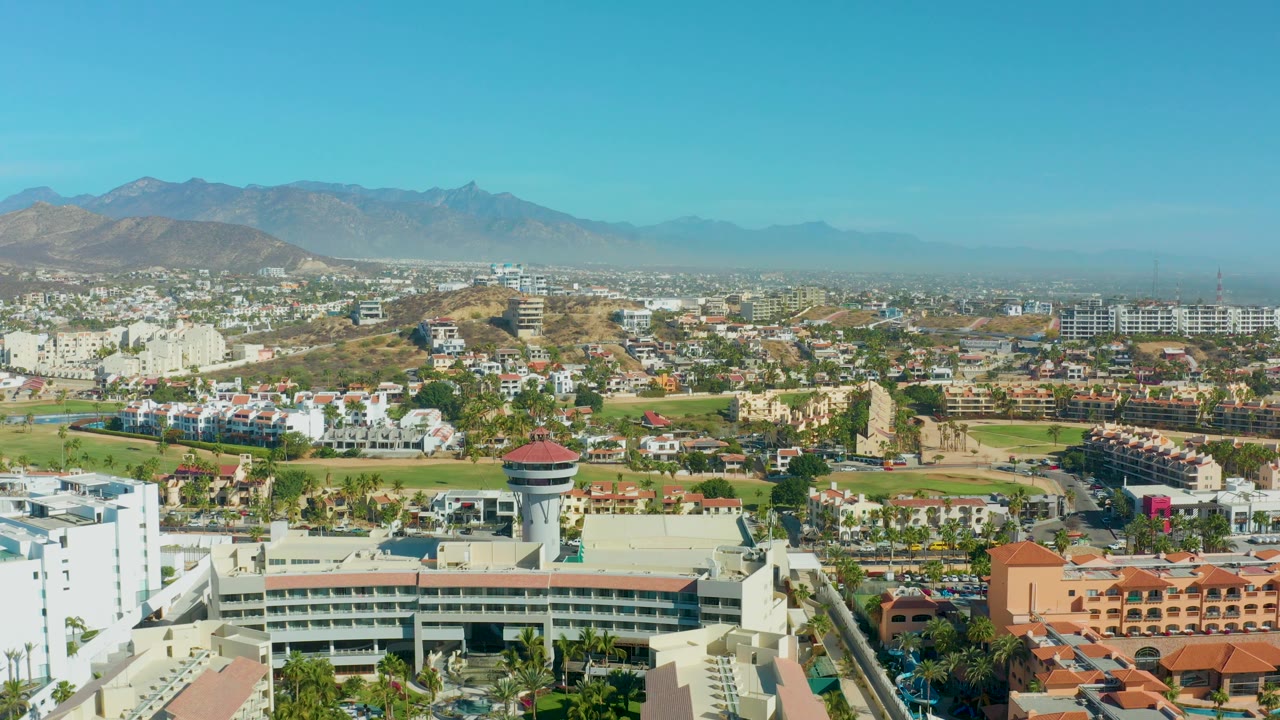 Wide urban aerial with white buildings, palms, dry hillside, and mountains