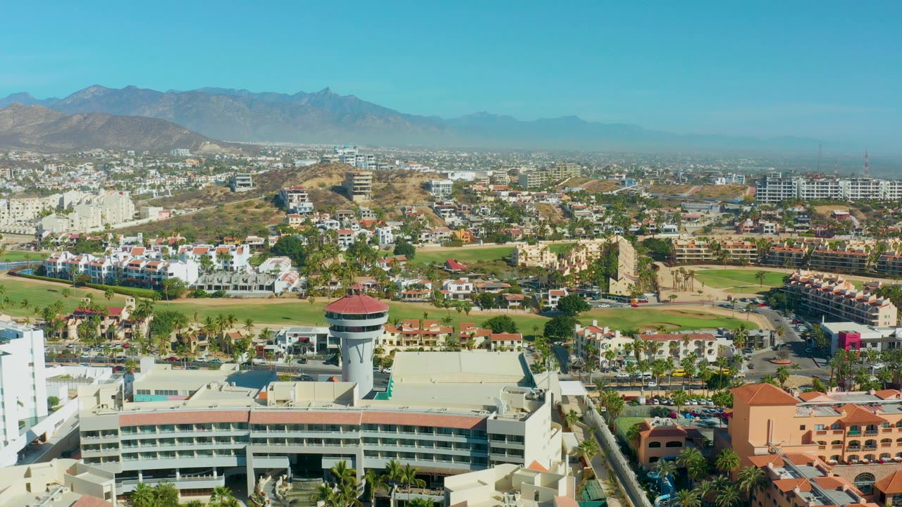 Aerial daytime view of San Jose del Cabo with a round-topped water tower in the lower center, white buildings, green open space, dry hills, distant mountains, blue sky, and no visible people.