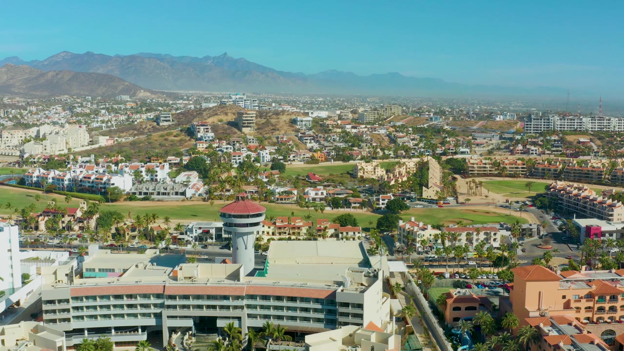 Wide San Jose del Cabo city panorama with the water tower below center, white residential clusters, a broad green open area, tan hills, and hazy mountain ridges in the background, with no visible people.