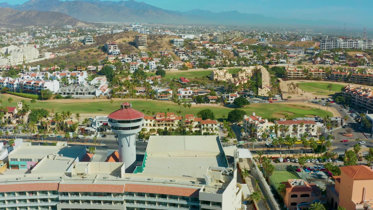 San Jose del Cabo aerial view one second in, with the water tower lower in frame, roads and parking areas near green fields, hillside housing, distant mountains, and no visible people.