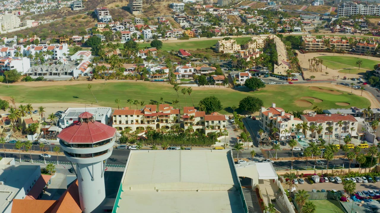 Aerial frame over San Jose Del Cabo with the red-roof control tower lower center-left, a pale roof filling the lower middle, parked cars, palm trees, residential buildings, green fairway, sand traps, hills, and no visible people.
