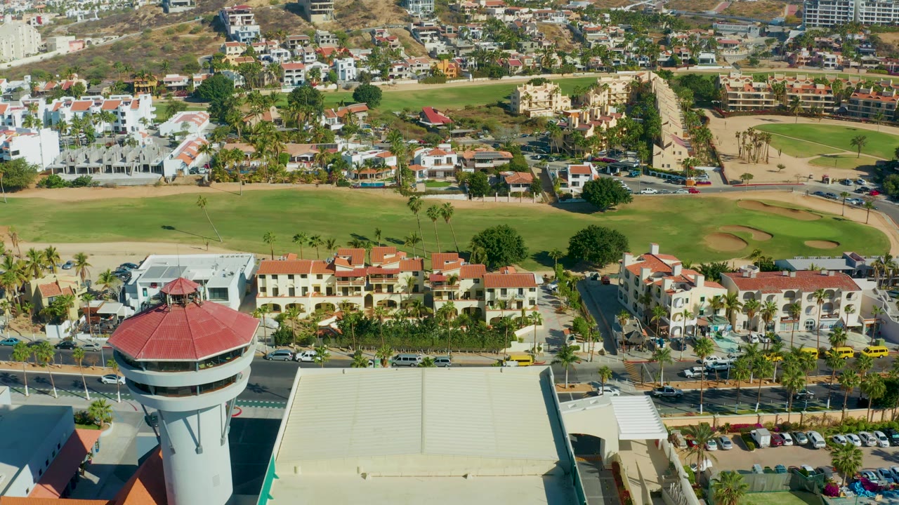 Aerial frame over San Jose Del Cabo with the control tower roof lower center, the large roof to the right, road traffic below, palm trees, homes, fairway grass, tan sand traps, hillside buildings, and no visible people.