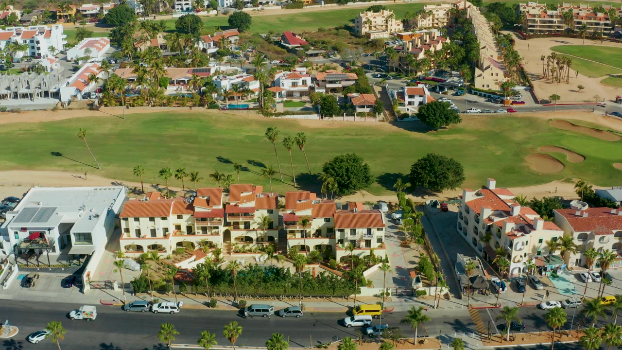 Opening aerial frame over San Jose Del Cabo residences with red tile roofs, a road with vehicles, green golf fairway, sand bunkers, palm trees, and no visible people.