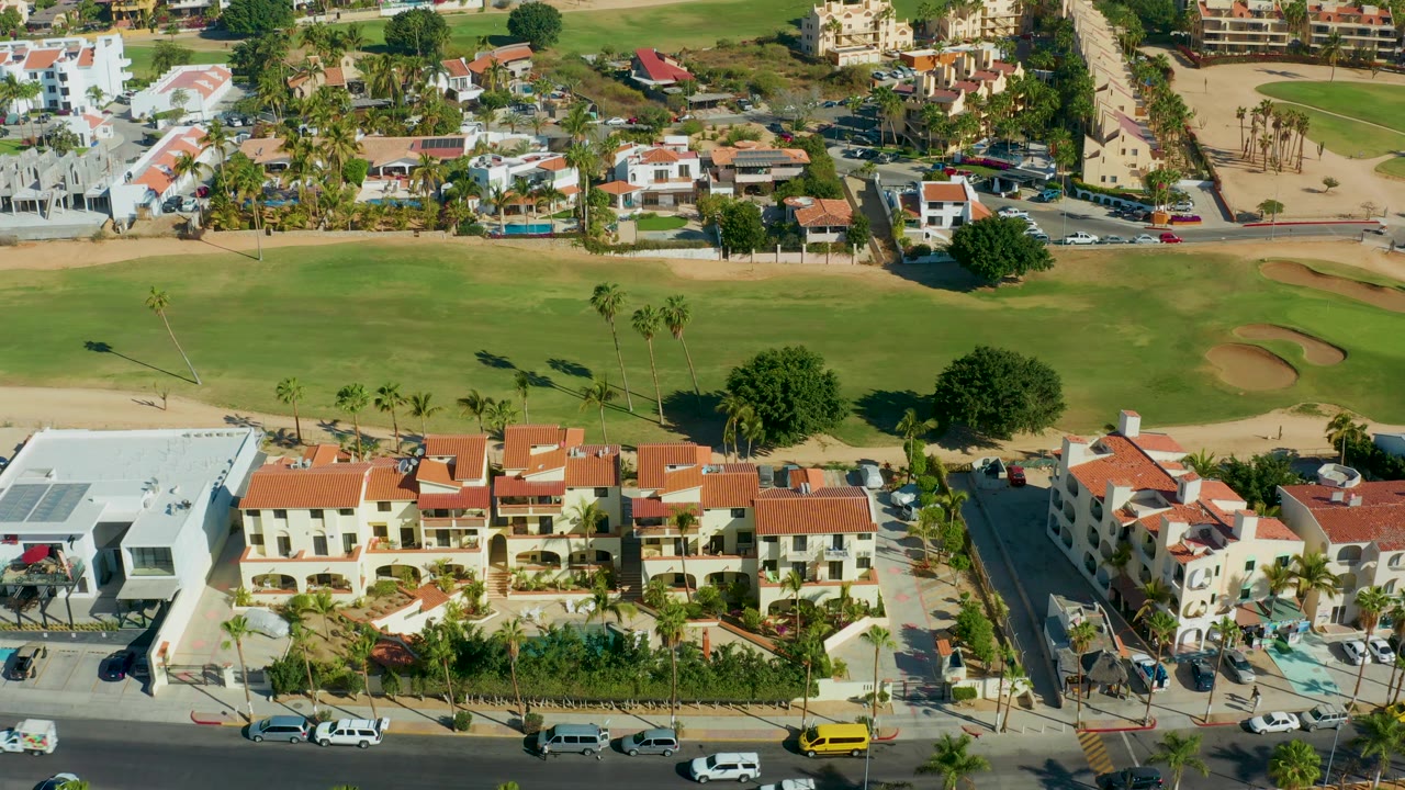 San Jose Del Cabo aerial frame with a white modern building, red-roof residences, road traffic below, green fairway and bunkers behind, and no visible people.