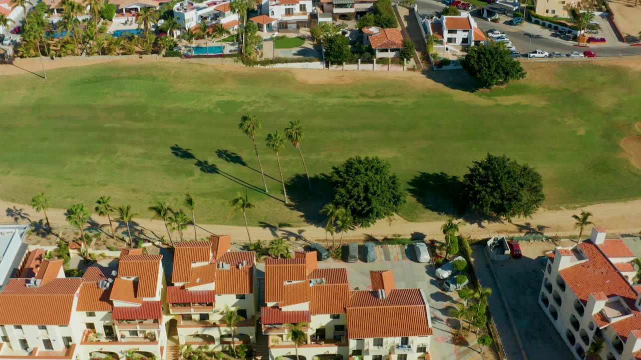 Aerial frame over San Jose del Cabo showing green golf course grass, palm trees, red tile roof homes, parked cars, and no visible people.