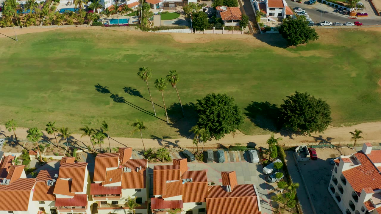 San Jose del Cabo golf course aerial with open grass in the center, palm tree shadows across the turf, homes at the lower edge, and no visible people.