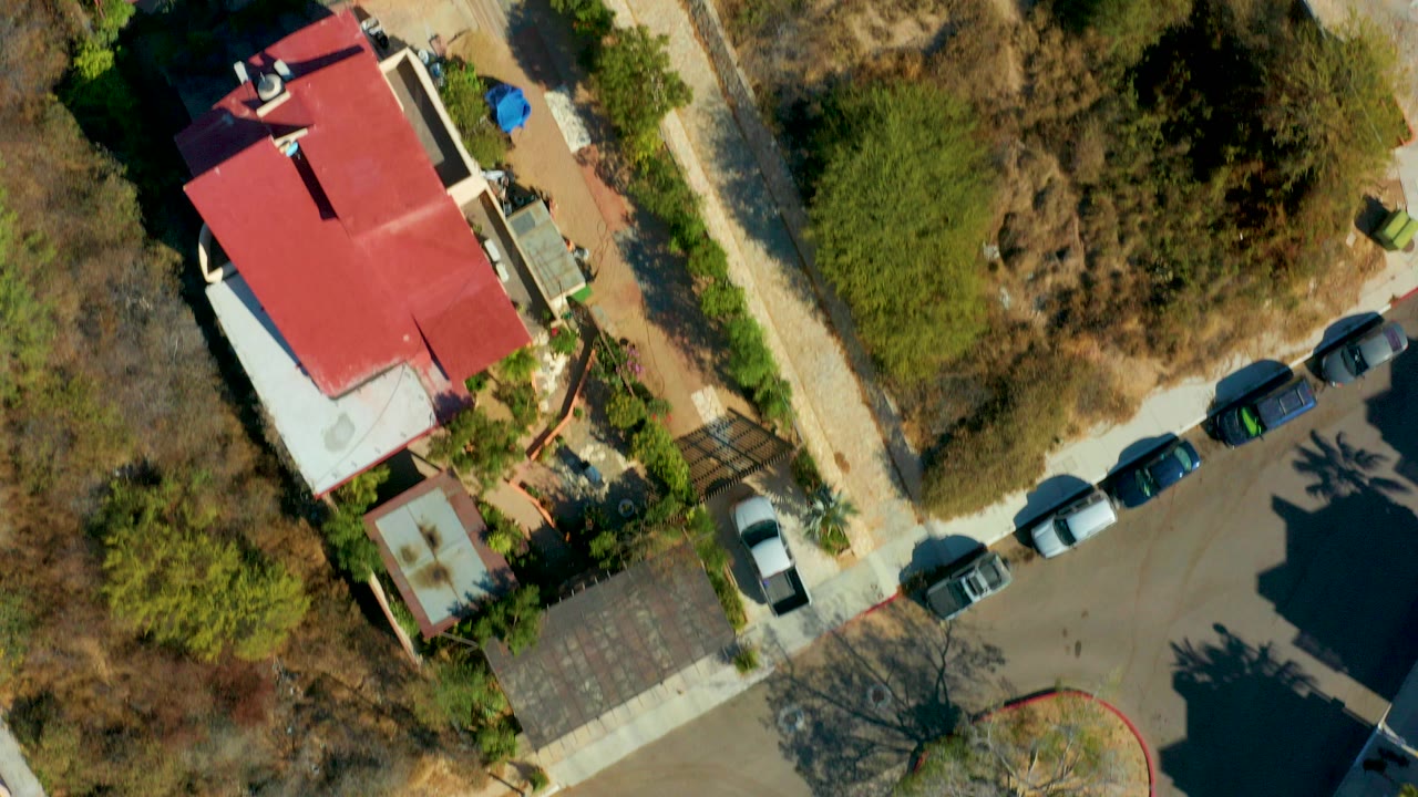 Overhead aerial frame in San Jose del Cabo showing a red-roofed building beside parked cars, a paved road, trees, and dry vegetation, with no visible people.