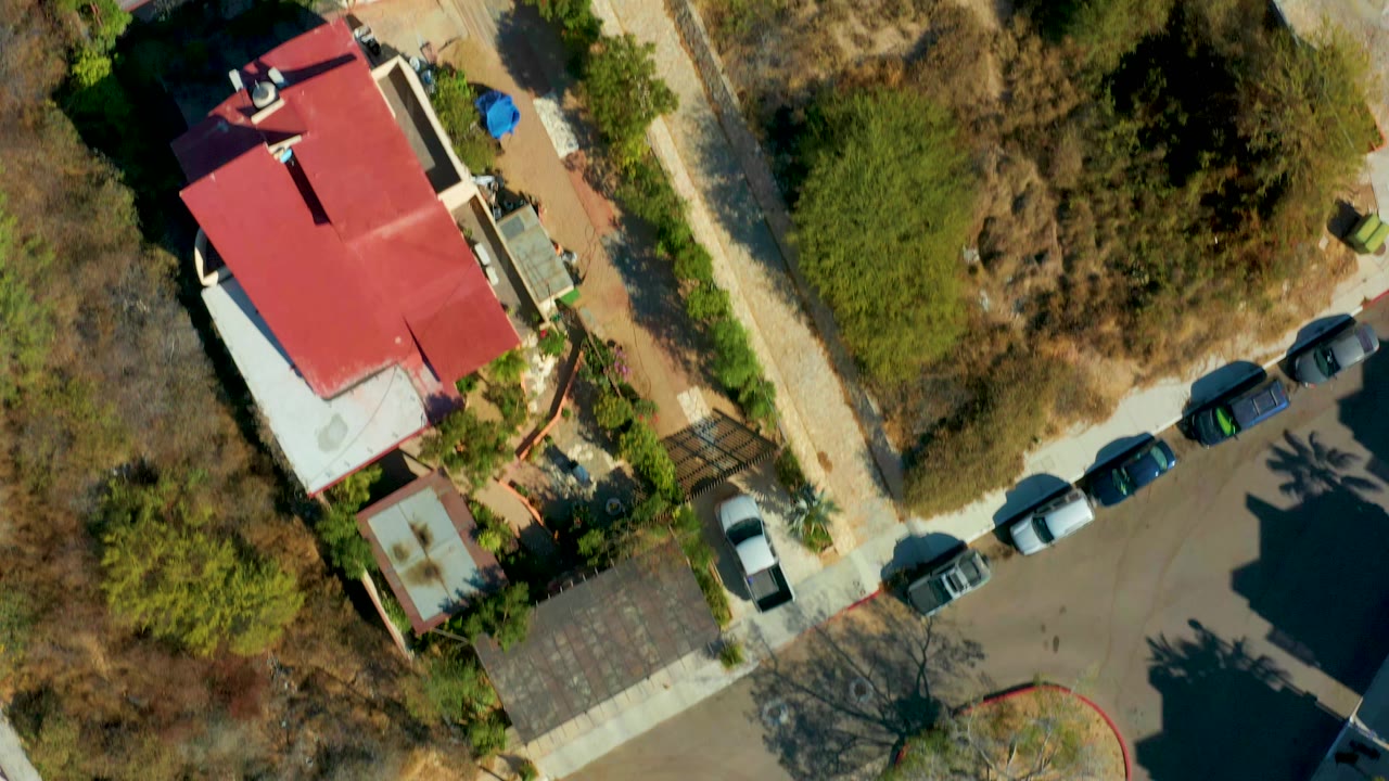 San Jose del Cabo aerial view holds over the red-roofed building while the dry lot, roadside parking, trees, and building shadows fill more of the frame, with no visible people.