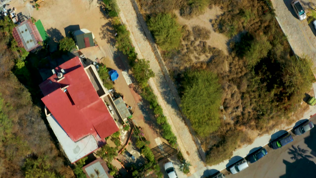 San Jose del Cabo top-down frame shifts the red-roofed building lower in the view, with roadside cars, dry brush, small yards, and service paths visible, with no visible people.