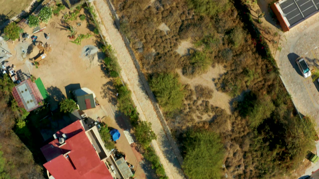Aerial view crossing dry vegetation, a red-roofed building corner, and a construction yard.