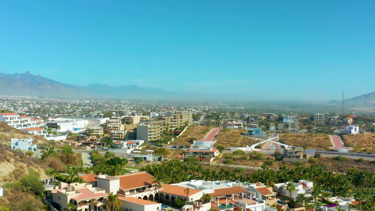 Wide aerial view of San Jose del Cabo with red-tiled roofs in the foreground, low-rise city blocks, dry hillsides, distant mountains, clear blue sky, and no visible people.