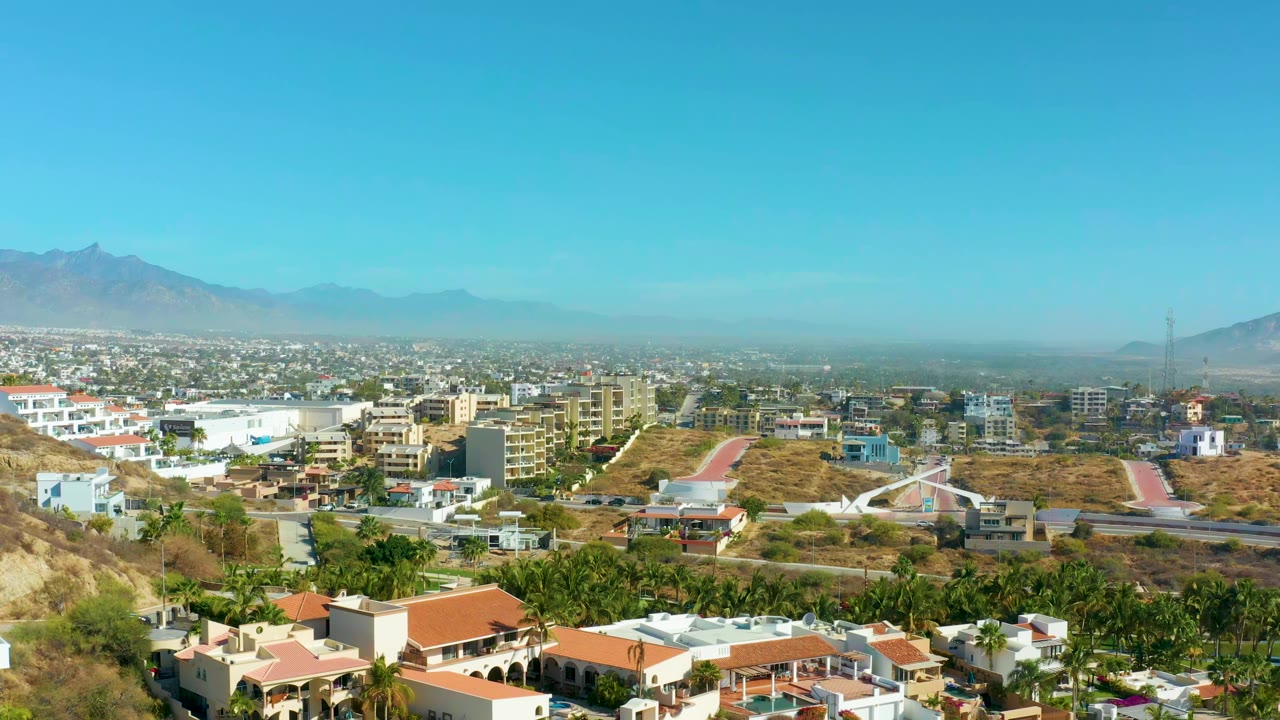 San Jose del Cabo aerial frame showing hillside villas and red roofs below, urban blocks across the middle distance, hazy mountains beyond, clear sky, and no visible people.