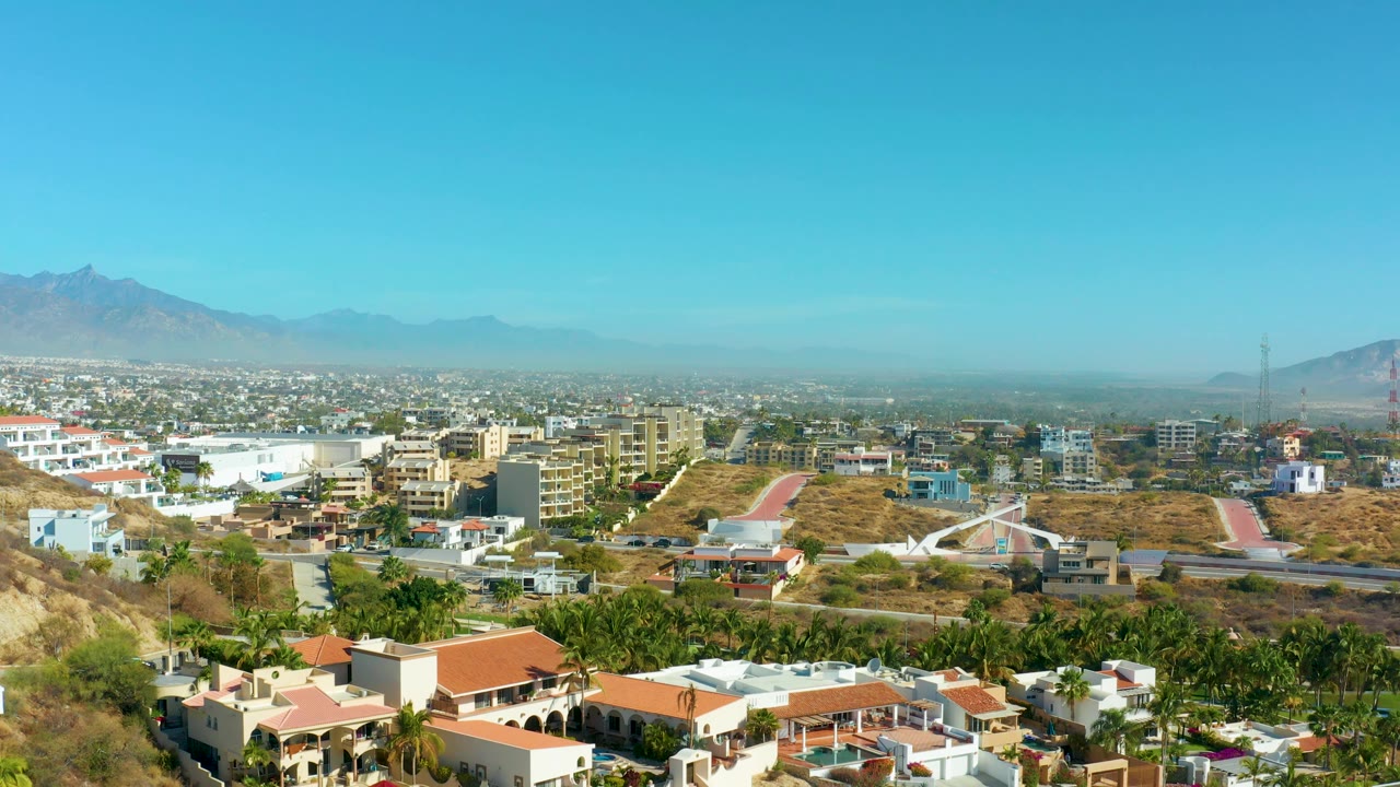 Elevated San Jose del Cabo view with white residential buildings and red roofs in front, roads and commercial-looking low-rise blocks in the center, mountains on the horizon, and no visible people.