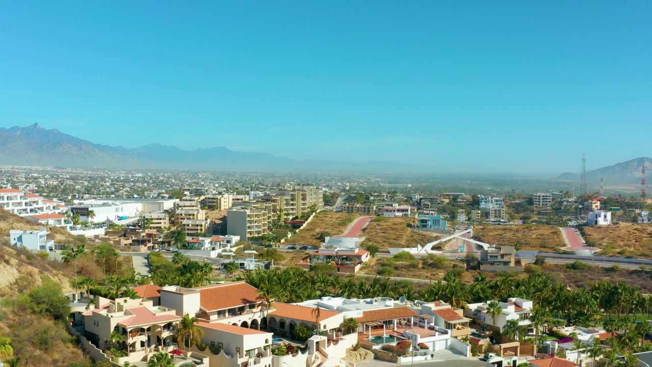 Cityscape across dry terrain with red-roofed houses and mountains under blue sky.