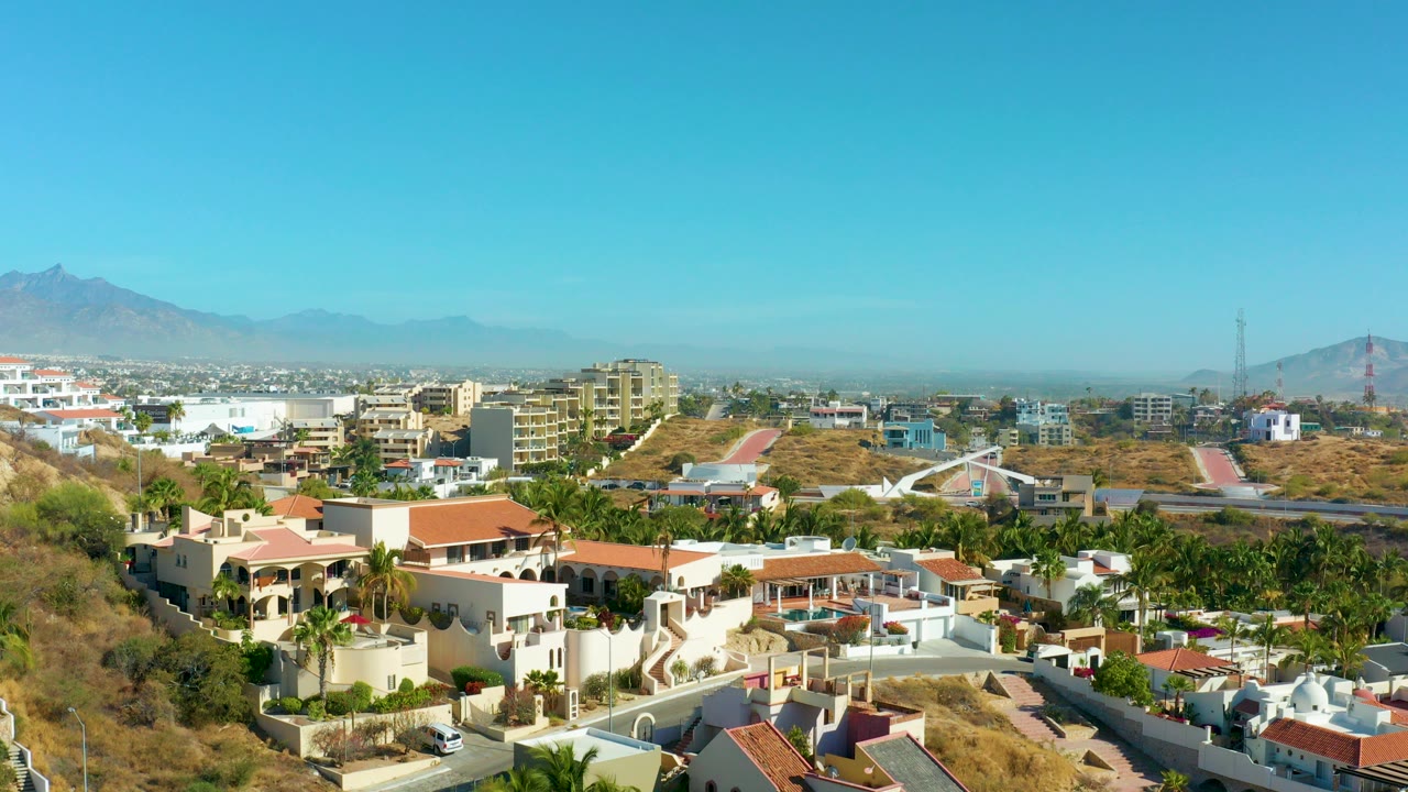 Wide elevated view of San Jose Del Cabo hillside homes with red-tiled roofs, palm trees, dry slopes, distant mountains, and no visible people.