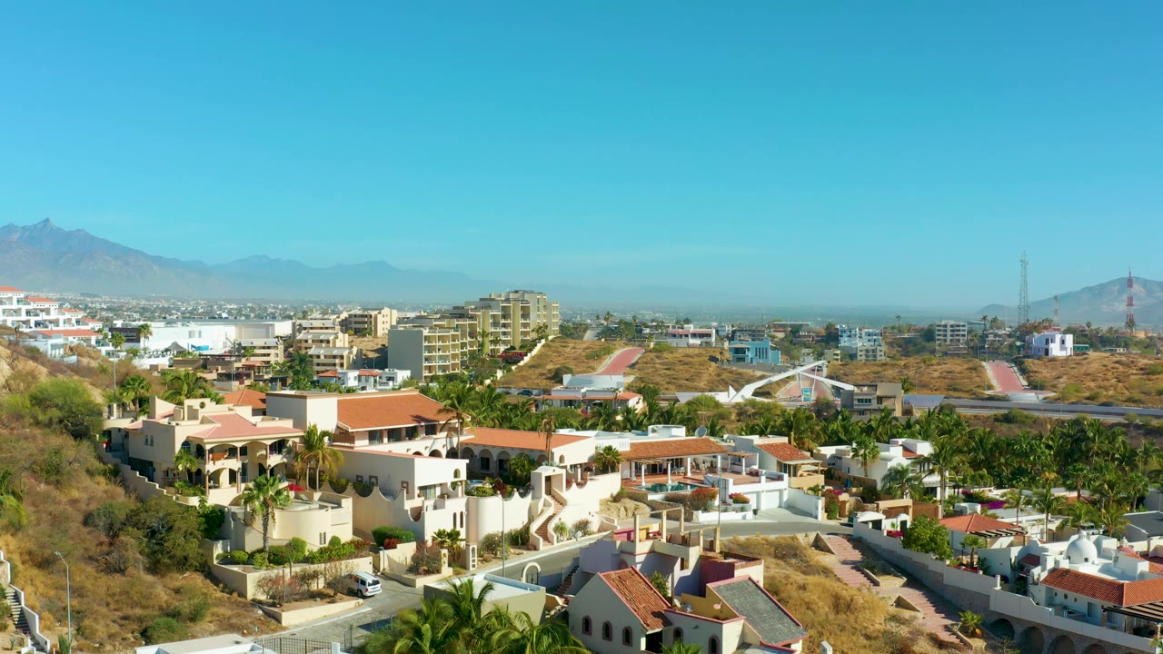 San Jose Del Cabo hillside neighborhood seen from above with red-roofed houses, palms, dry lots, midrise buildings, mountain silhouettes, and no visible people.