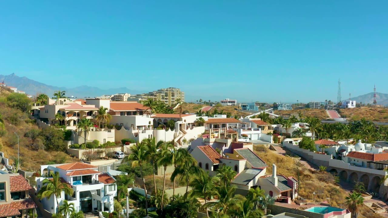 San Jose del Cabo residential hillside seen from above, with white villas, red roofs, palms, dry terrain, distant buildings, and no visible people.