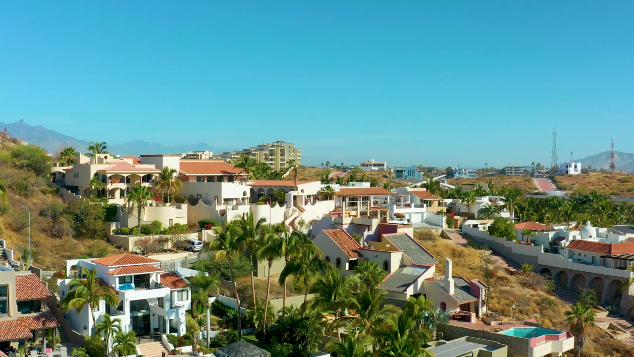 Aerial frame of San Jose del Cabo hillside housing with palms in the foreground, white homes and red roofs across the slope, and no visible people.