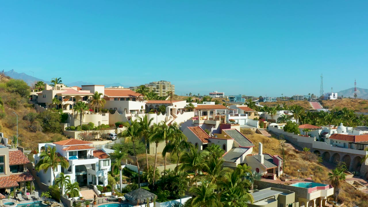 White hillside homes, palms, pools, and dry terrain beneath a clear sky.