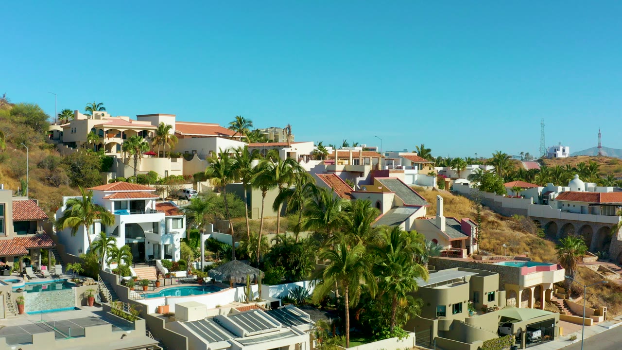 Elevated San Jose del Cabo residential view with white and beige hillside homes, red tile roofs, palm trees, pool terraces, dry slope, and no visible people.