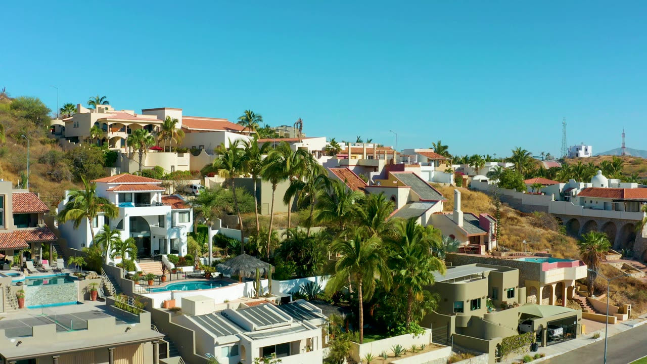 San Jose del Cabo residential hillside with tall palms in front of modern homes, a blue pool, road sections, dry terrain, and no visible people.