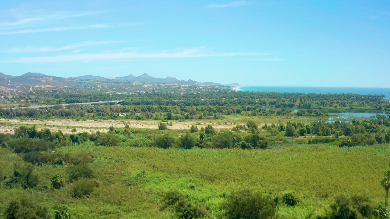 San Jose del Cabo coastal plain with a broad green field in the foreground, a pale road across dry ground, water areas to the right, distant hills, ocean, and no visible people.