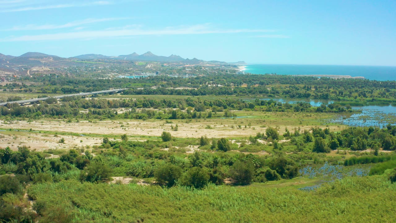 Early frame of the San Jose del Cabo coastal landscape showing green foreground vegetation, pale dry fields, a road, blue water, distant mountains, ocean, clear sky, and no visible people.