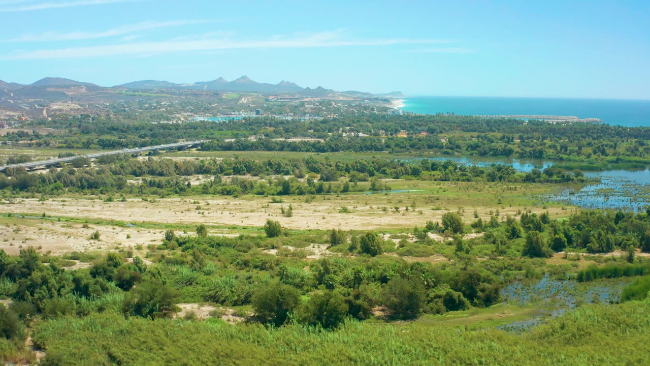 At 0:02, the San Jose del Cabo frame shows more blue wetland patches at right, sandy fields below, a road through the trees, distant hills, ocean, coastline, and no visible people.