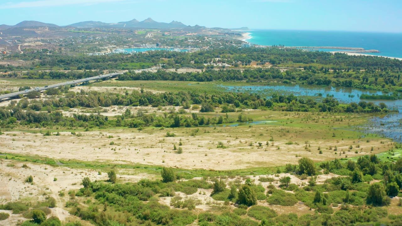 Wide aerial frame of San Jose del Cabo showing pale dunes across the lower frame, wetland water and trees through the center, a road running beside the river, ocean at upper right, mountains at upper left, and no visible people.