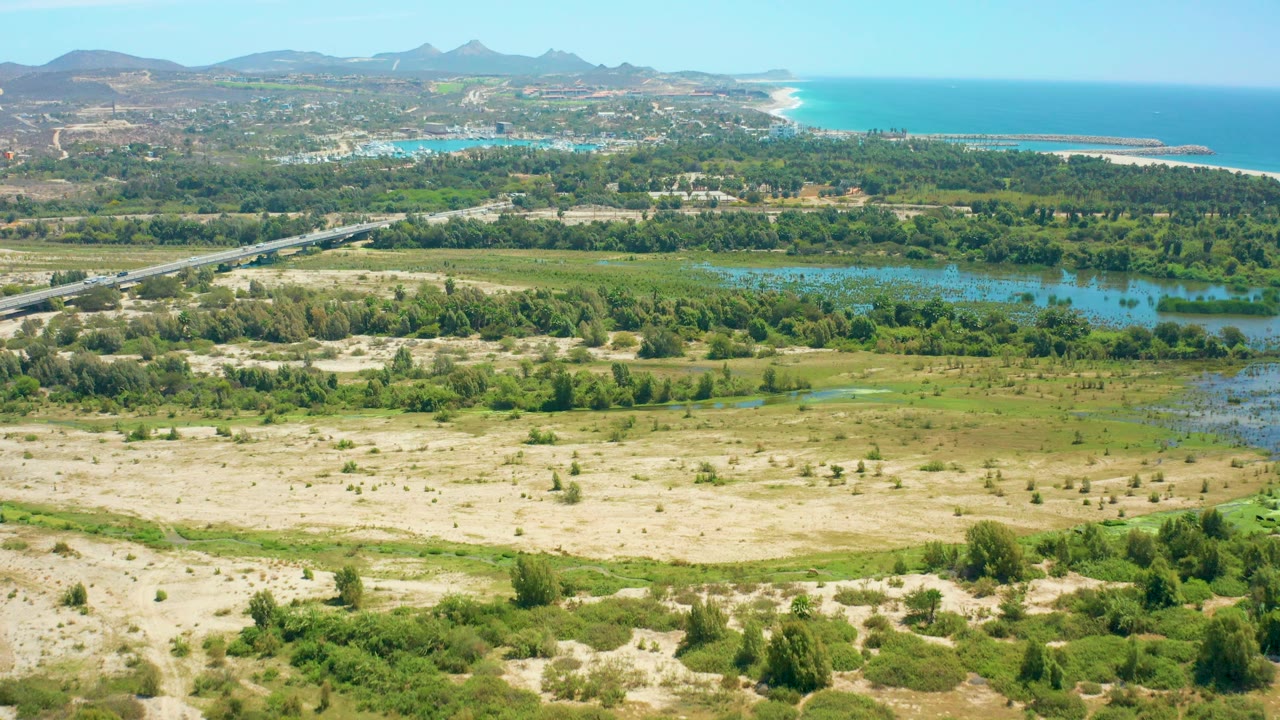 Elevated view in San Jose del Cabo with sandy terrain and low shrubs below, a green river valley and road crossing the center, a curved coastline and bright blue ocean behind, mountains in the distance, and no visible people.