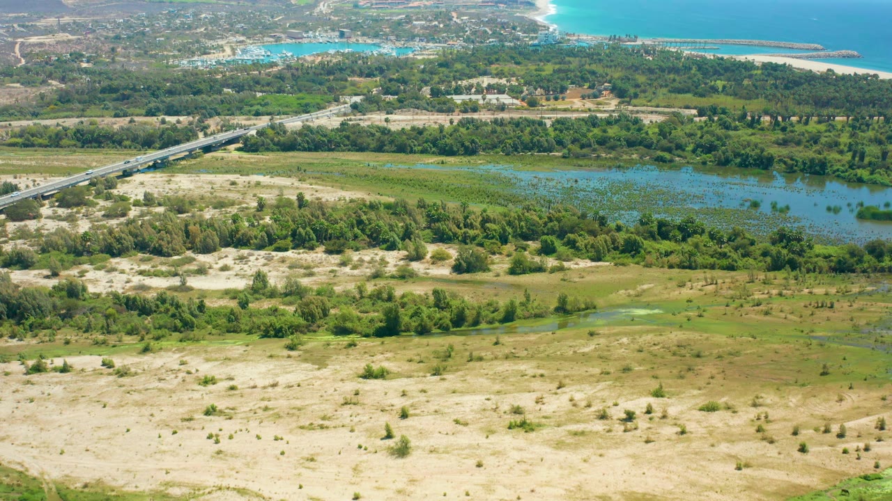Aerial frame over San Jose Del Cabo with wetland water spread across the midground, green tree lines around it, sandy terrain below, and no visible people.