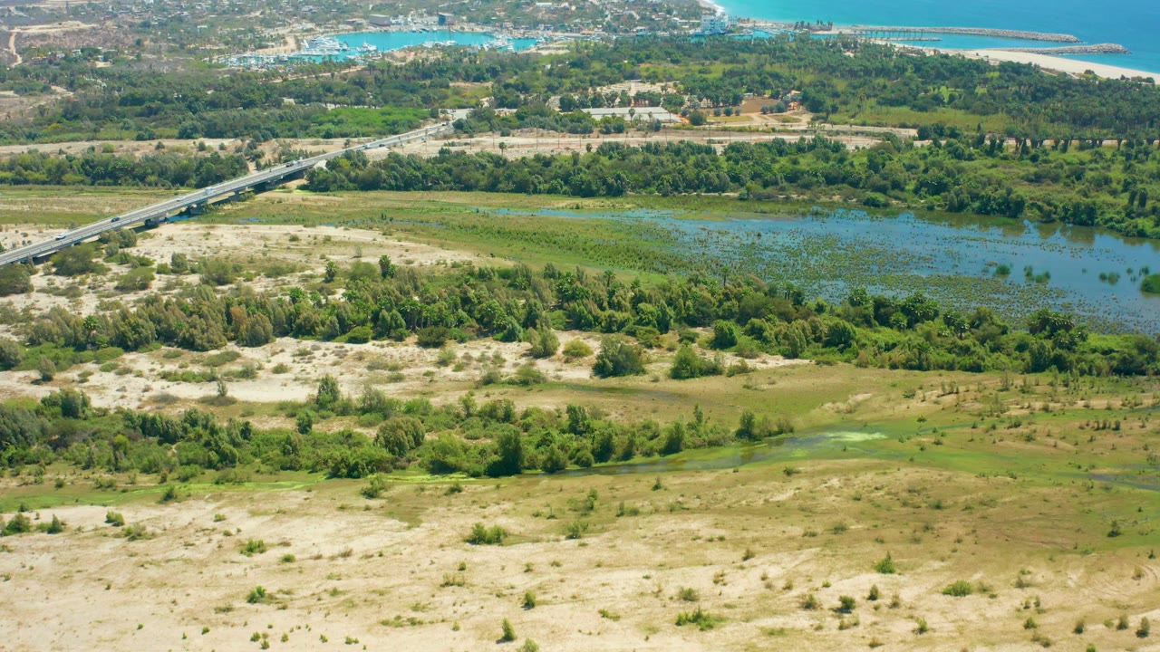 Aerial frame over San Jose Del Cabo with the bridge more prominent on the left, shallow blue-green wetland water centered, sandy patches below, and no visible people.