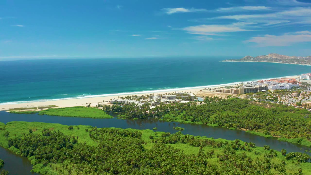 Opening aerial frame over San Jose Del Cabo with a winding river in green vegetation, a pale beach, blue ocean, distant hills, scattered clouds, and no visible people.