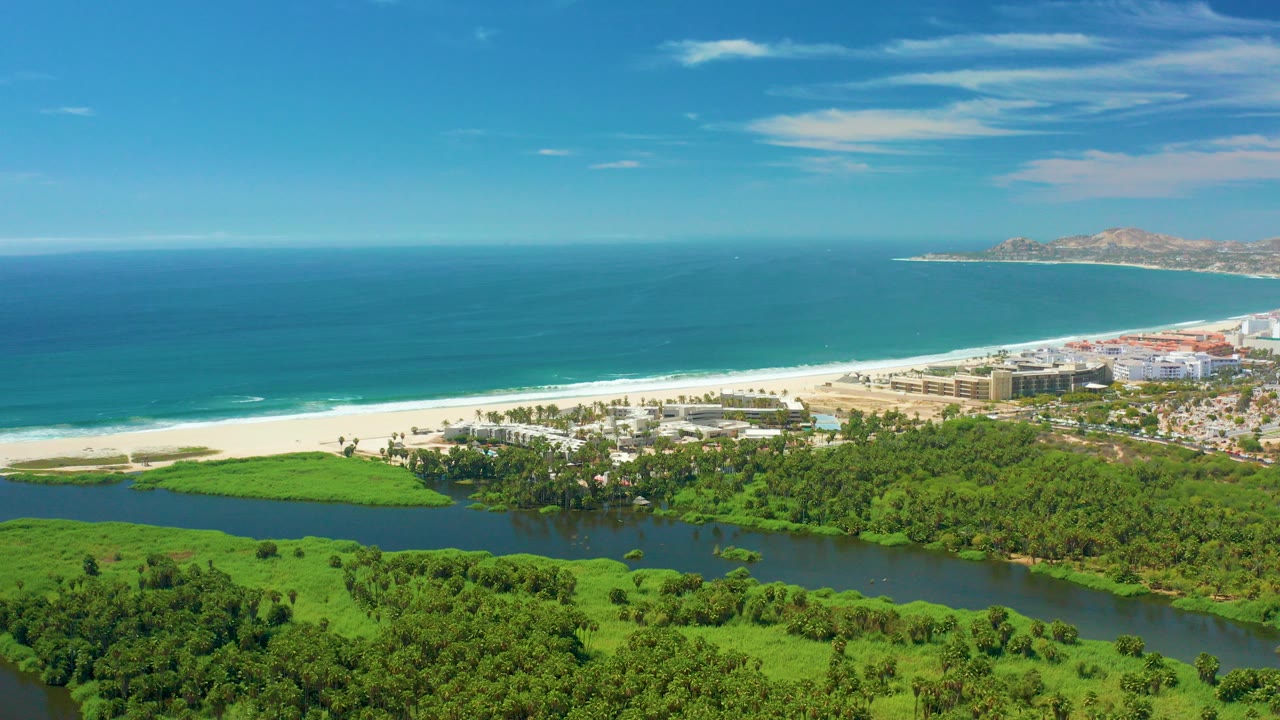 Aerial view over San Jose del Cabo showing green wetlands in front, a dark lagoon, white beach, blue ocean, coastal buildings, distant headland, and no visible people.