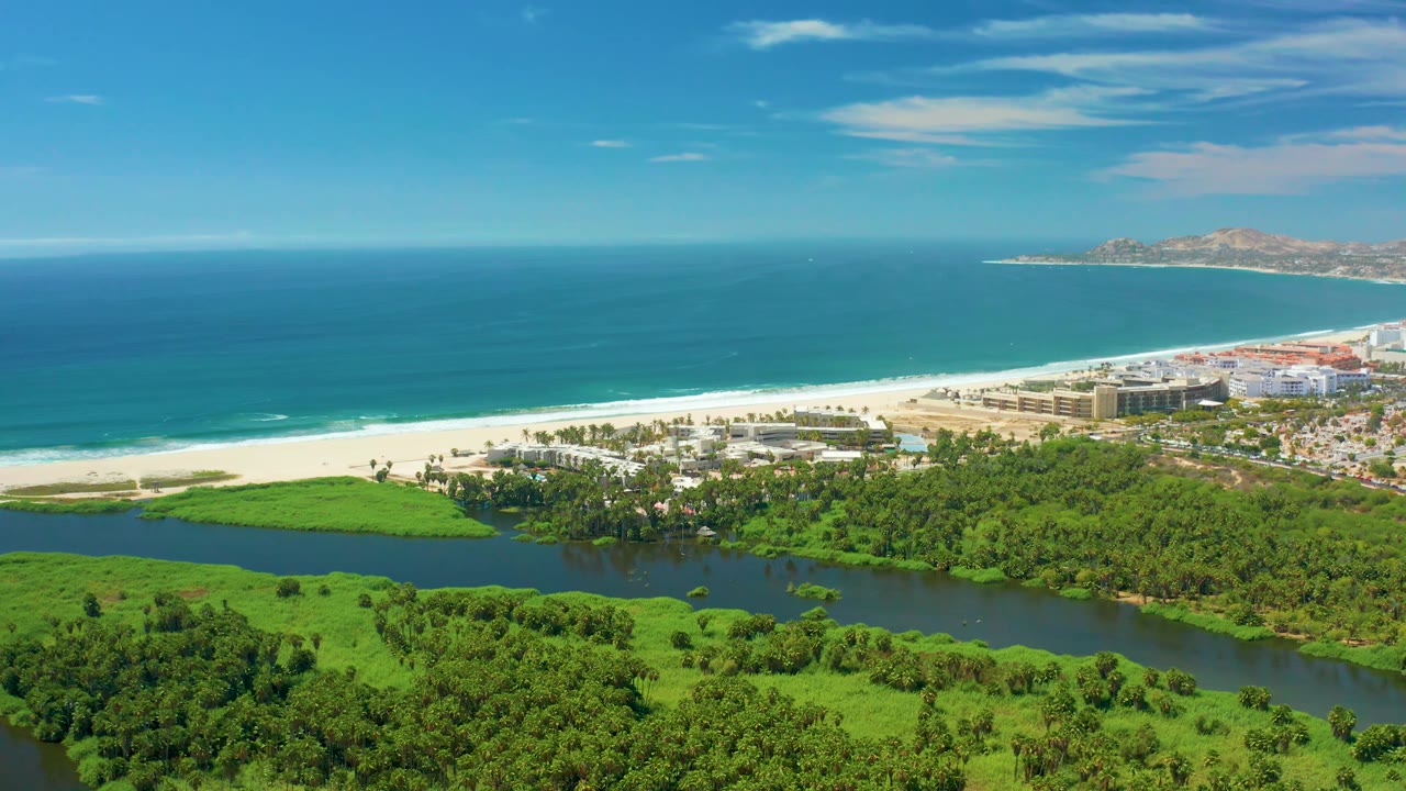 Aerial view in San Jose del Cabo with bright beach and ocean spanning the middle and background, green wetland water in the foreground, buildings on the right edge, and no visible people.