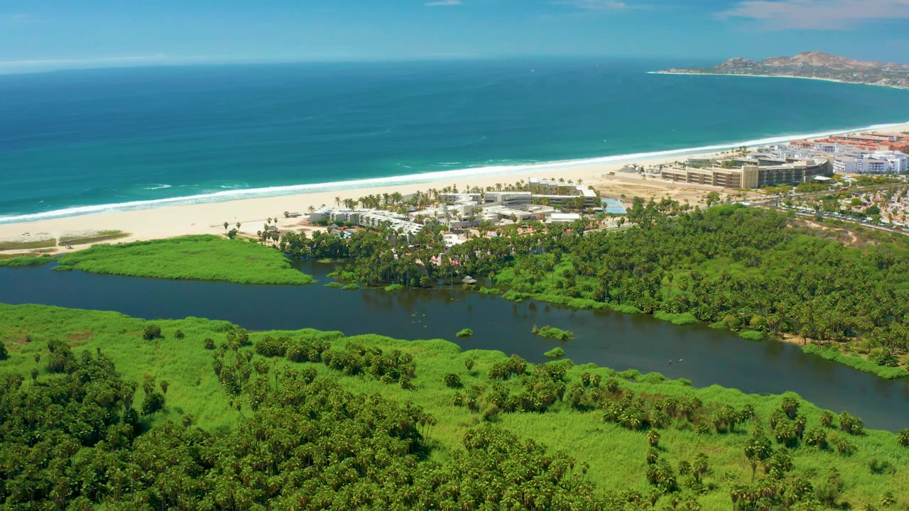 San Jose del Cabo shoreline seen from above, with the dark lagoon curving through green vegetation, white beach and ocean beyond, buildings at the right side of the coast, and no visible people.