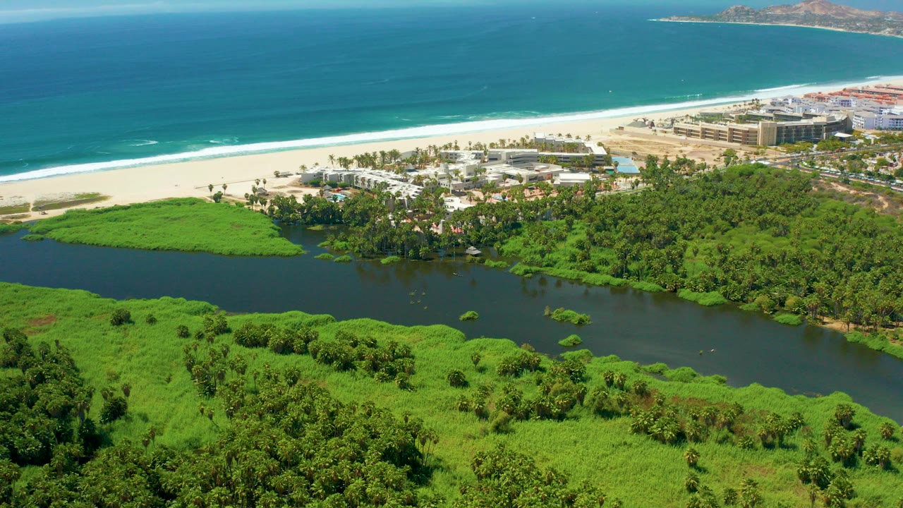 Aerial frame over San Jose del Cabo showing a dark river bordered by dense green vegetation, a white sand beach, coastal buildings, blue ocean, and no visible people.