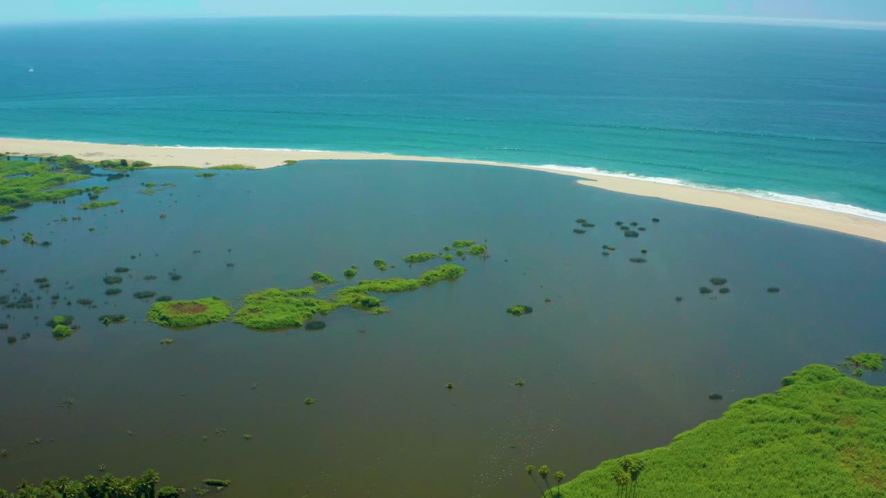Aerial frame over San Jose del Cabo showing calm lagoon water with scattered green vegetation, a pale sand beach, turquoise ocean, white surf, and no visible people.