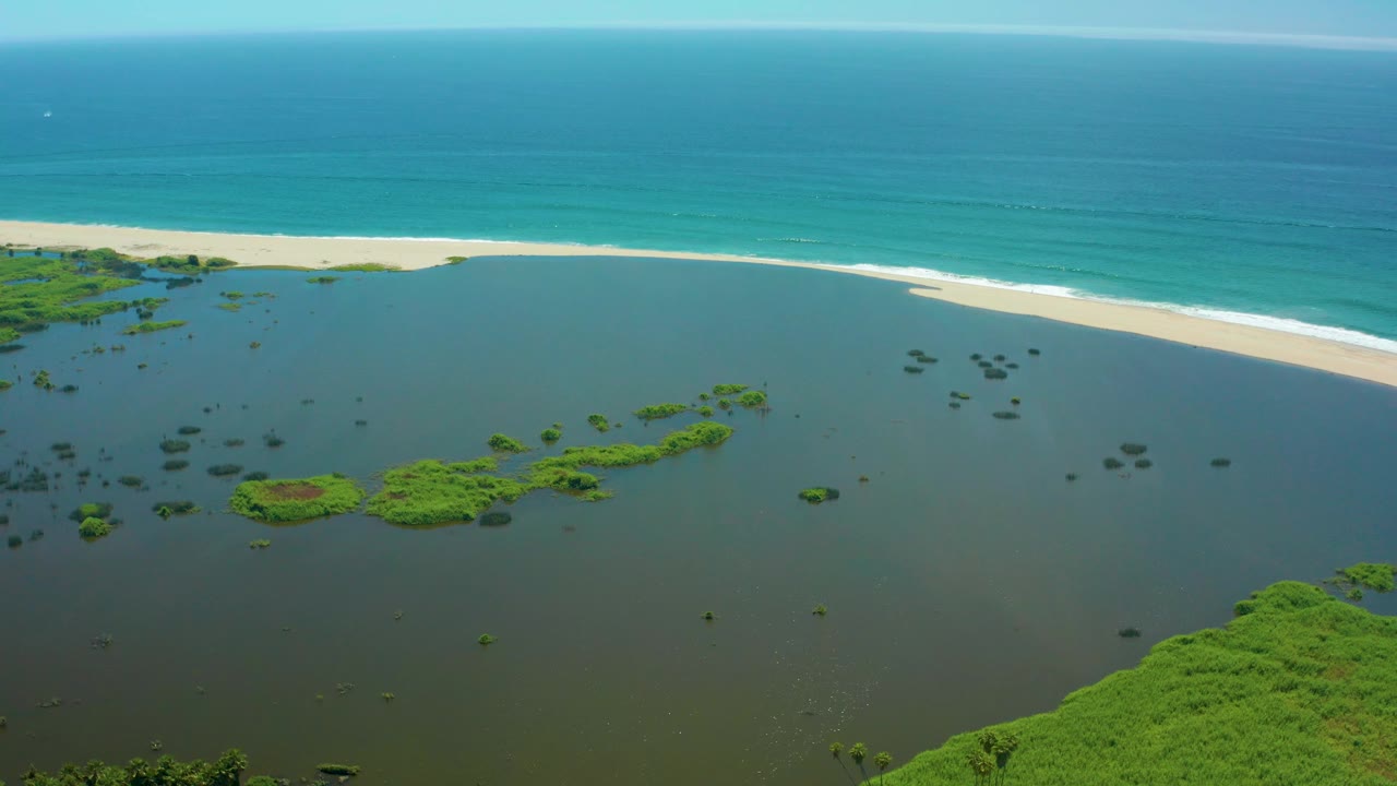San Jose del Cabo aerial frame with calm lagoon water in the foreground, scattered green vegetation, a bright sandy beach, white surf, blue ocean, and no visible people.