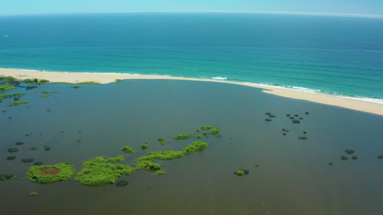 Aerial view in San Jose Del Cabo showing shallow lagoon water with green vegetation below, a pale sandy beach across the middle, turquoise ocean beyond, and no visible people.