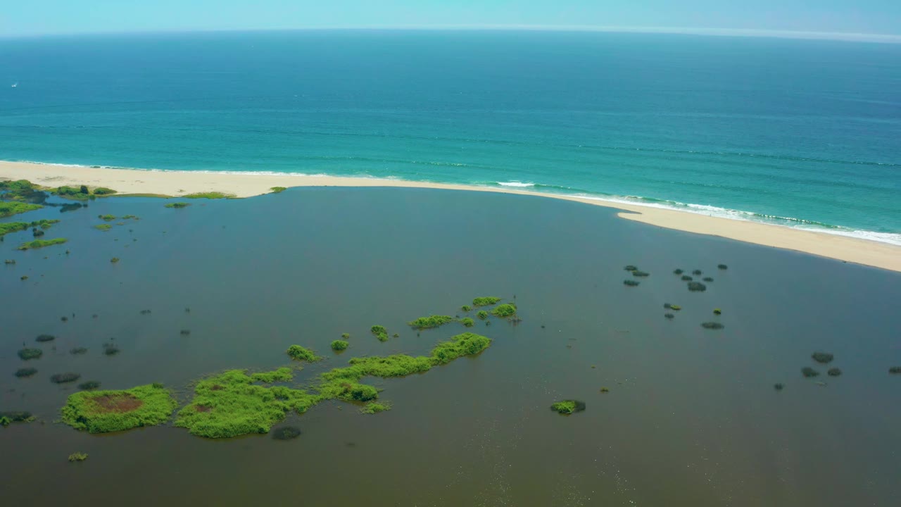 San Jose Del Cabo coastal lagoon remains in the foreground while the sand beach and thin white surf line stretch diagonally across the frame, with no visible people.