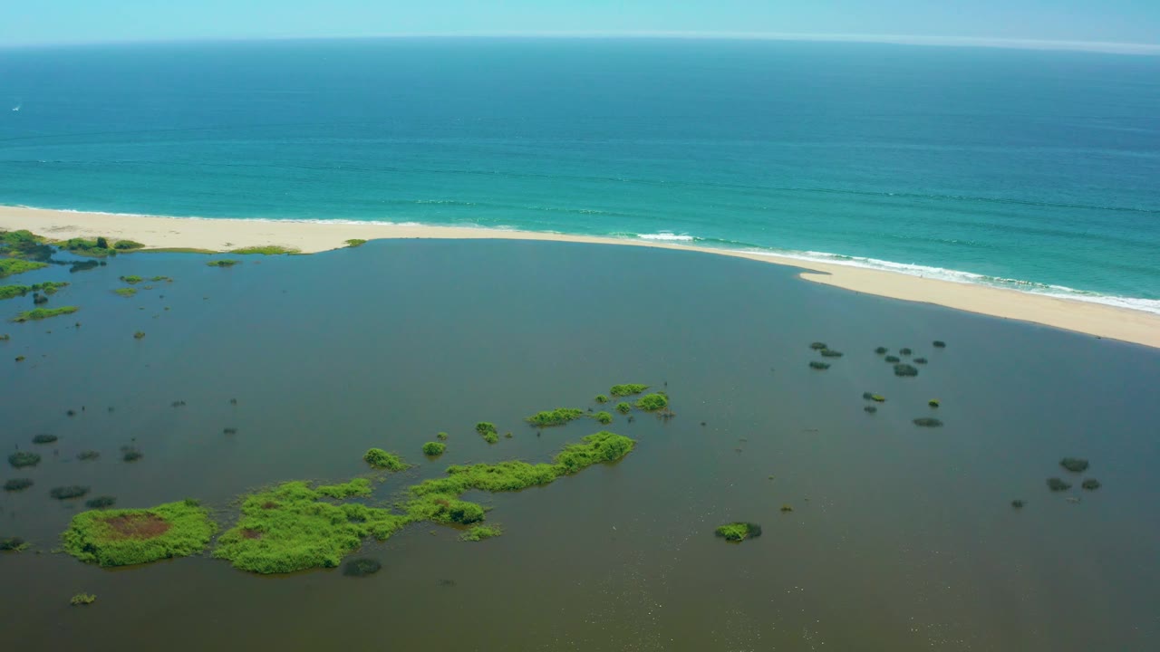 Wide aerial frame over San Jose Del Cabo coastline shows dark green lagoon shallows, clustered vegetation, a continuous sandy beach, blue ocean bands, and no visible people.