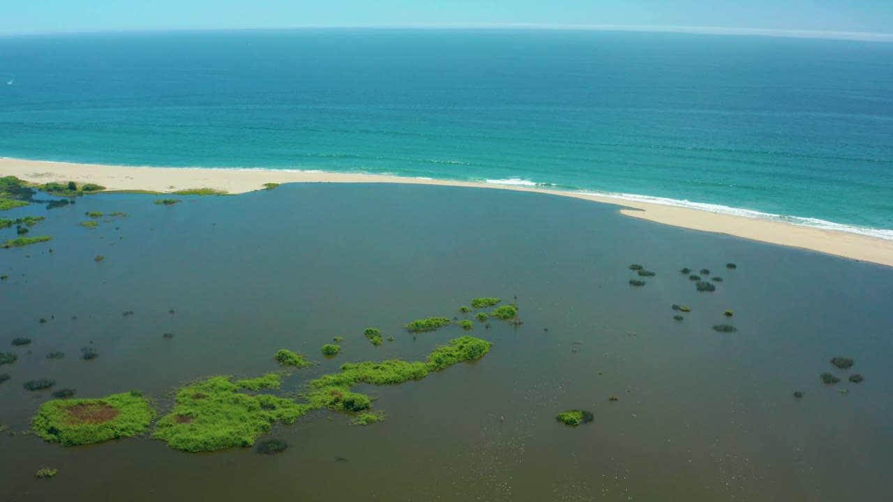 Aerial view in San Jose Del Cabo keeps the lagoon low in frame with scattered vegetation, the beach running left to right, calm turquoise ocean above, and no visible people.