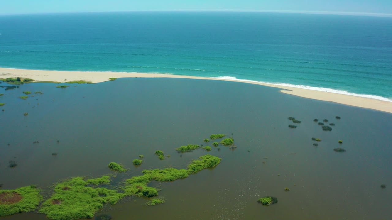 Aerial frame over San Jose del Cabo showing shallow lagoon water with green vegetation patches, a pale sand strip, turquoise ocean, clear sky, and no visible people.