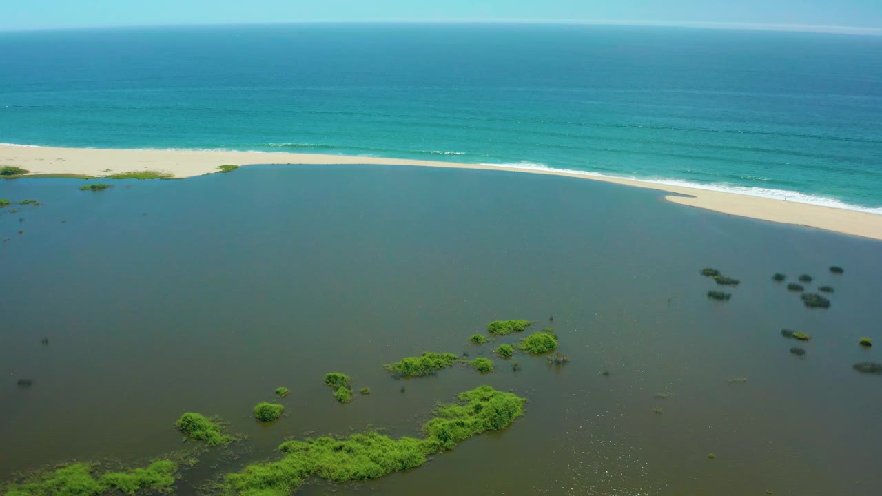High aerial frame from San Jose Del Cabo with scattered green vegetation in dark lagoon water, a narrow sand strip at the shoreline, turquoise ocean, clear sky, and no visible people.