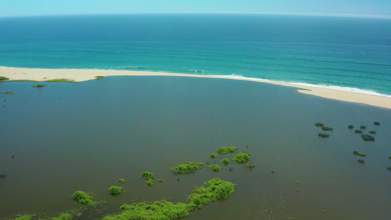 San Jose Del Cabo aerial still with green vegetation clustered near the lower frame, dark lagoon water in the foreground, a bright sand beach, white wave line, and no visible people.