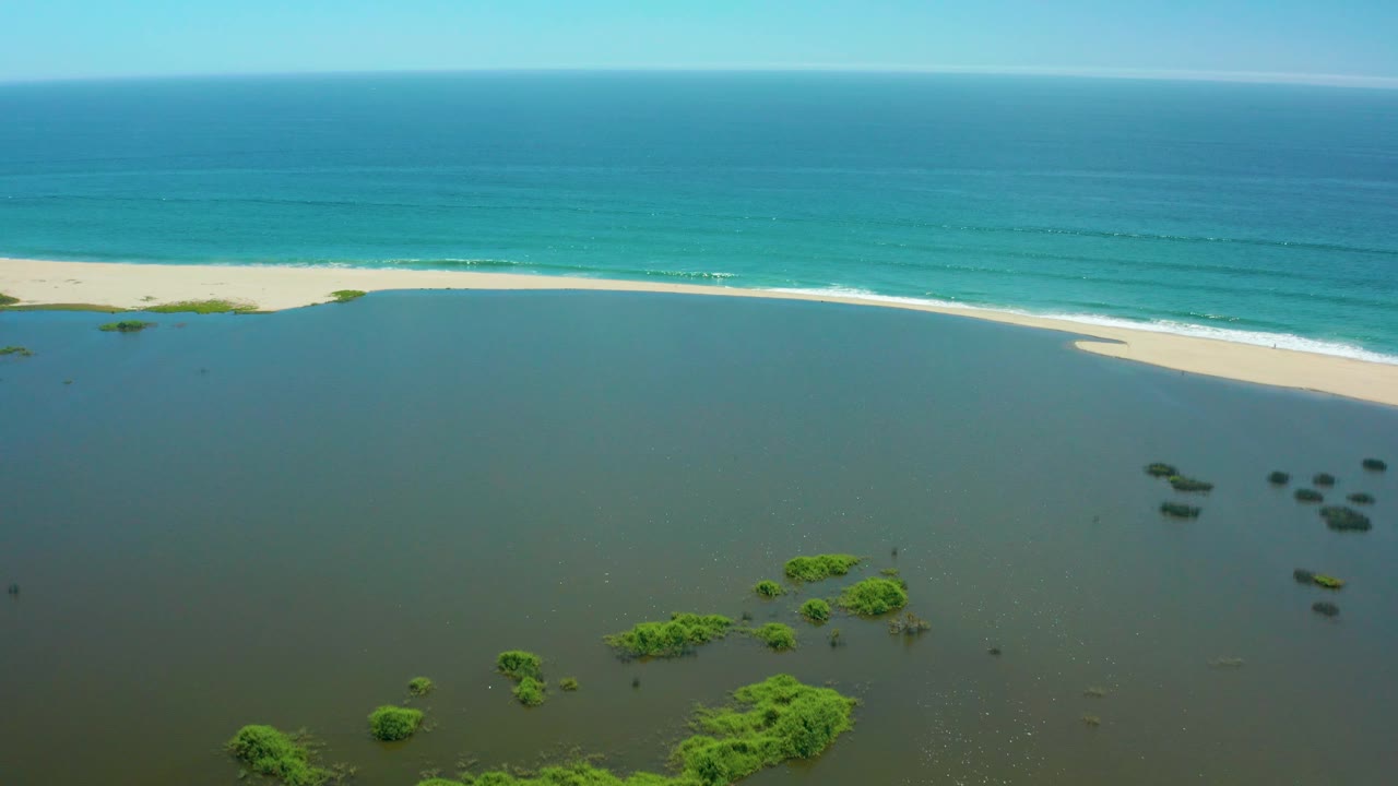 Wide San Jose Del Cabo coastal frame showing open lagoon water in front, a light sandy beach and foamy surf across the middle, layered turquoise ocean behind, and no visible people.